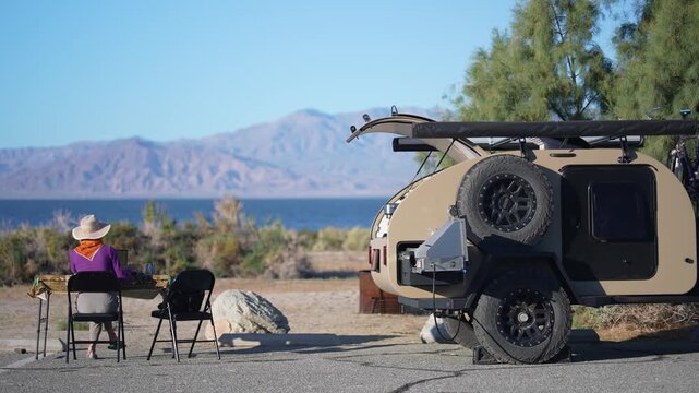 Mature woman sits at table working on computer next to her teardrop camper trailer by the beach at Salton Sea in California.