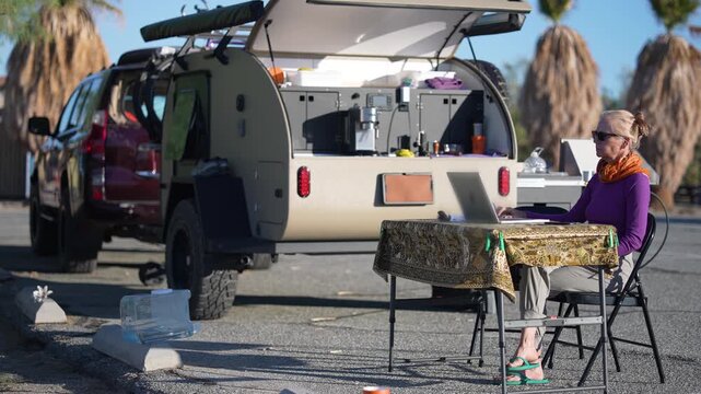 Mature woman sits at a table working on a computer by her teardrop trailer by the beach in Californias Salton Sea area