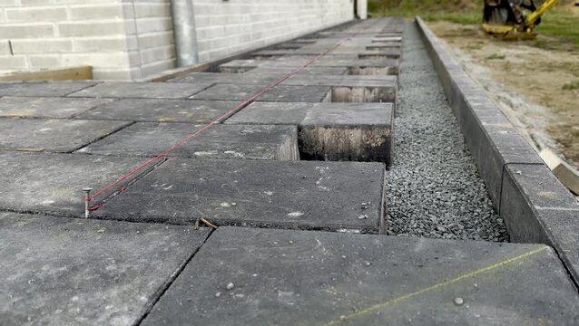 Construction site showing black paving stones being laid in a straight line with gravel base and red string line for alignment near a building exterior