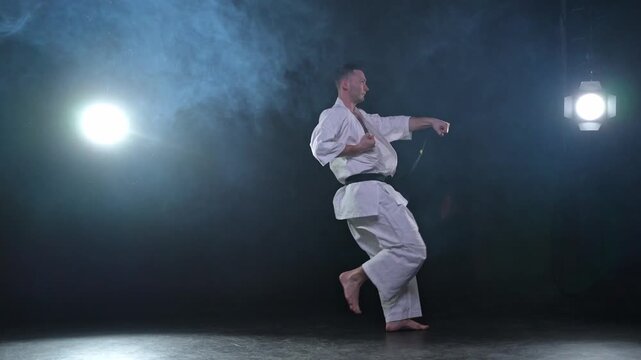 Strong man fighter in white kimono for karate martial arts with black belt preparing for fight training in black background. Man training moves punches kicks practicing shadow fight ready for sparring