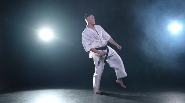 Strong man fighter in white kimono for karate martial arts with black belt preparing for fight training in black background. Man training moves punches kicks practicing shadow fight ready for sparring