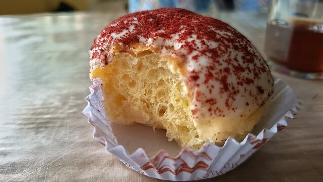 Close-up of a soft vanilla cupcake topped with white icing and red velvet crumbs, served in a paper liner on a marble table. Sweet bakery dessert with shallow depth of field and blurred tea backgroun