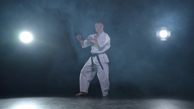 Strong man fighter in white kimono for karate martial arts with black belt preparing for fight training in black background. Man training moves punches kicks practicing shadow fight ready for sparring