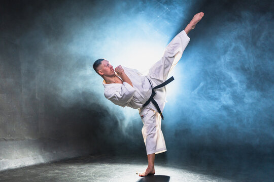 Strong man fighter in white kimono for karate martial arts with black belt preparing for fight training in black background. Man training moves punches kicks practicing shadow fight ready for sparring