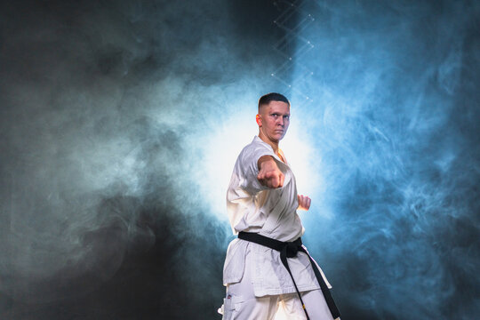 Strong man fighter in white kimono for karate martial arts with black belt preparing for fight training in black background. Man training moves punches kicks practicing shadow fight ready for sparring