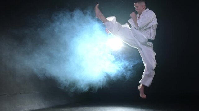Strong man fighter in white kimono for karate martial arts with black belt preparing for fight training in black background. Man training moves punches kicks practicing shadow fight ready for sparring