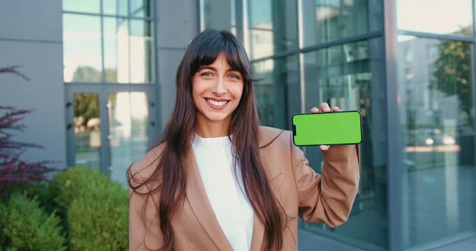Confident businesswoman in suit smiling and holding a smartphone with a green screen. Perfect for showcasing mobile apps, user interface and UX designs, software demos, or tech startup presentations