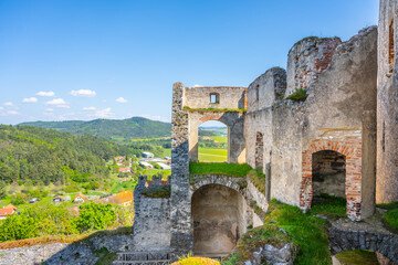 Obraz premium Rabi Castle stands as a large ruin in Bohemia. Visitors can see the stone walls and views of the village below. The sky is clear, and the landscape shows green hills and houses.