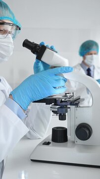 Female scientist wearing a mask and blue safety gear, using a microscope for research while a male colleague works in the background. Medicine, healthcare and science concept