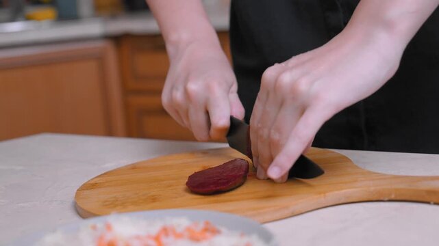 Close-up of hands slicing boiled beets on the table, home cook trimming bright red root with a sharp knife on a wooden cutting board, bowl of grated carrots visible, calm atmosphere of kitchen prepara