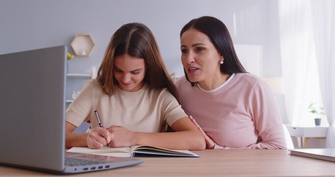 Portrait of school girl sitting at table in front of laptop, doing homework with mother helping her. Happy parent and child cooperating on tasks, discussing difficult questions while studying online.
