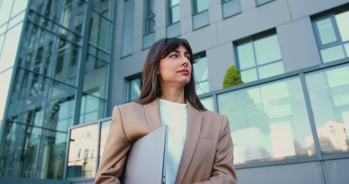 Professional business woman with long dark hair holding a laptop and standing in a trendy co working space, looking at the away with a friendly smile, outdoors