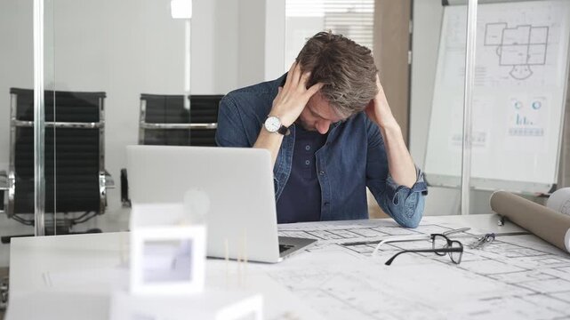 Stressed architect feeling overwhelmed and frustrated, suffering from burnout while working on blueprints for a tight project deadline in a modern office setup