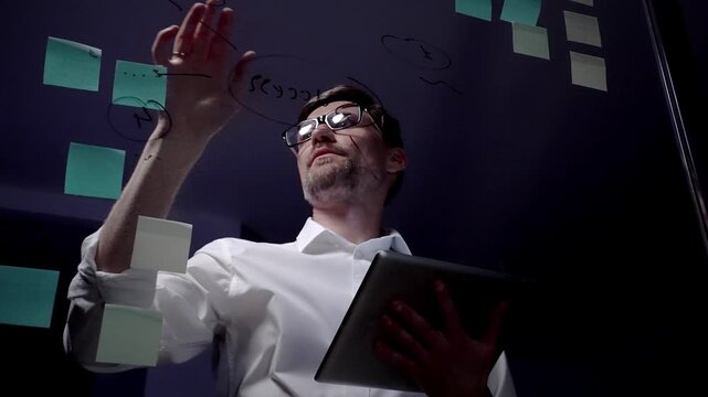 Man in eyeglasses organizing thoughts and ideas for business optimization on a clear glass board. Creating a strategic roadmap for innovation and project planning