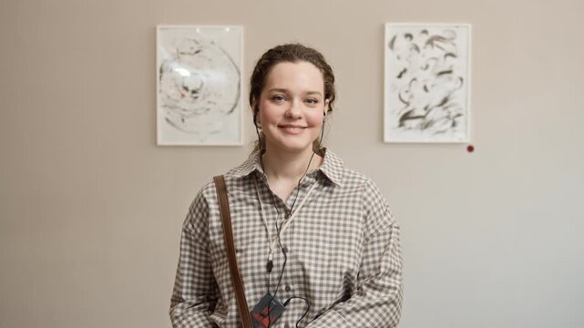 Zoom in slowmo portrait of young Caucasian woman wearing audio guide earphones looking at camera in modern art gallery with framed abstract paintings in blurred background