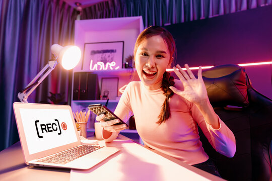 Host channel of young beautiful Asian looking at camera with social media online for greeting with fanclub holding smartphone, using laptop screen to present at neon colorful modern studio. Stratagem.