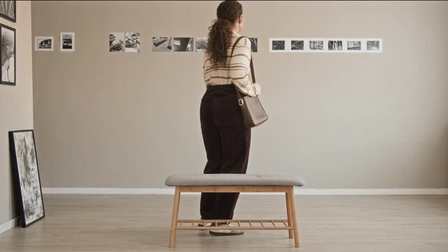 Full stab shot of young Caucasian female gallery visitor in casualwear listening audio guide in earphones taking seat in front of wall to observe modern black and white photography exhibition