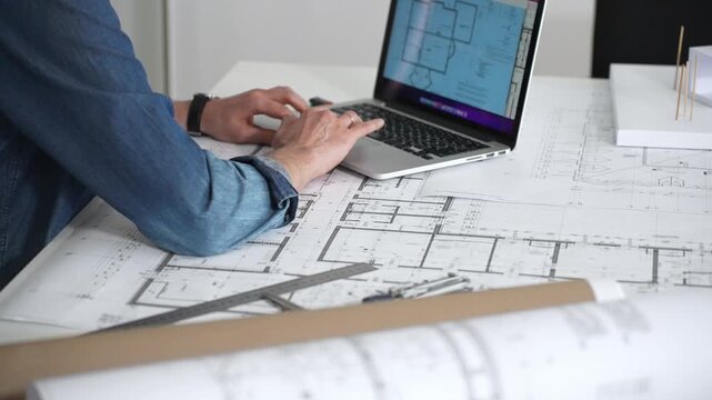 Architect sketching building plans on paper using a ruler and pen while working digitally on a laptop with a blueprint software displaying a floor plan
