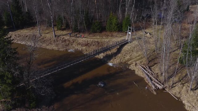 Aerial scene in Latvia shows a wooden suspension footbridge, timber gateway, driftwood, boulder, and walkers on a dirt path as brown water flows in warm early spring light.
