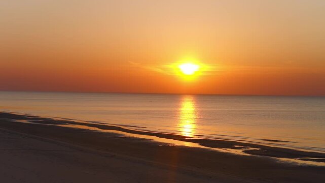 Aerial sunset over the Baltic Sea coast of Latvia shows a low sun, glassy water, sandbars, tidal pools, seabirds, slow drone pan, soft waves, and warm tones.