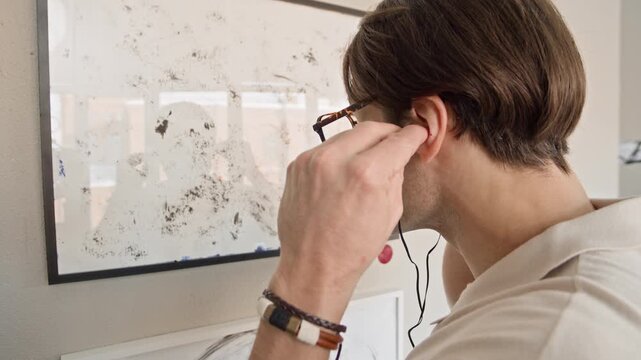 Handheld tilt up shot of young Caucasian man in eyeglasses standing in front of exhibition wall with abstract painting and using audio guide device, putting on wired earphones