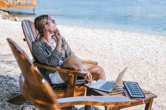 Young woman working on laptop by the sea with portable solar charger, digital nomad and workation lifestyle, remote work, sustainable travel and beach office concept.