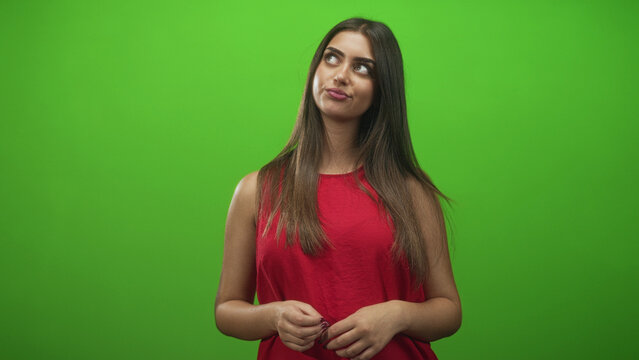 Young hispanic woman with hands clasping and slight head tilt, looking upward and fiddling fingers in a green screen studio; thoughtful reflection.