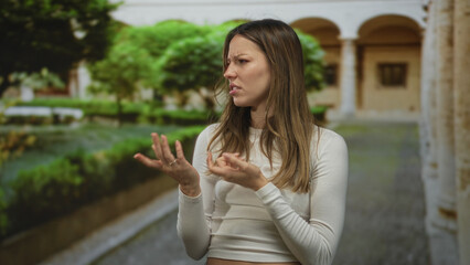 Fototapeta premium Woman holding her thumb and gesturing with both hands while wearing a white long sleeve crop top in an old town building; frustration confusion.