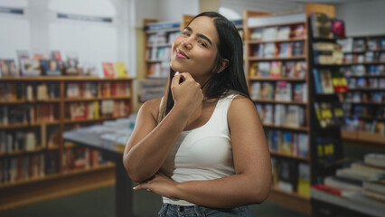 Woman touching chin with hand while smiling inside bookstore building wearing white tanktop and...