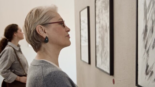 Arc slowmo shot of focused mature Caucasian woman in glasses observing framed abstract art painting with another young female gallery visitor viewing exhibition in background