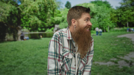Fototapeta premium Young caucasian bearded man in plaid shirt shrugs shoulders in green park under bright sunlight with blurred trees; confusion.