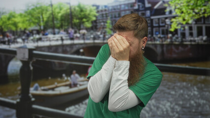 Man wearing volunteer shirt and long beard covers face with hands on street canal bridge under city...