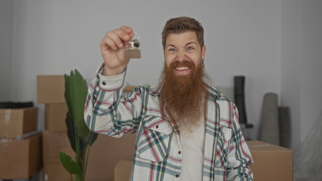 Man with beard holds house key and grins amid stacked moving boxes inside new building foyer; excitement.