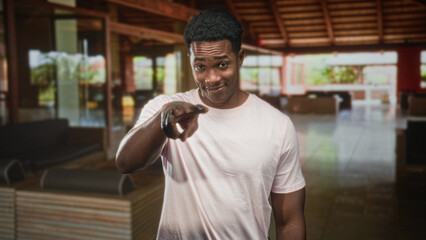 Obraz premium African american man in pale pink tshirt smiles at camera inside a hotel building lobby; friendly welcome.