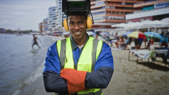 Man welder in welding helmet, reflective vest and gloves smiling with arms crossed on a sandy beach by a building; confidence skill duty.