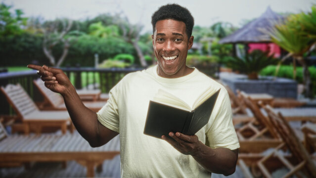 Man holding book and pointing finger on building deck, smiling while showing open pages and raised hand; casual happiness.