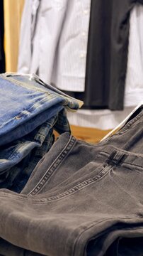 Folded denim jeans stacked on table with hanging shirts in background, warm wood shelving and visible price tag, stylist arranging casual menswear display for everyday shoppers, cozy retail atmosphere