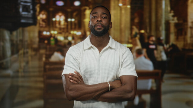 Man with arms crossed showing forearms and a bracelet in an ornate catholic church building, pews and chandeliers visible; quiet contemplation.