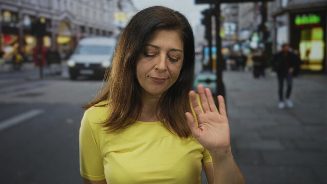 Middle aged hispanic woman waves hand amidst a bustling crowd of pedestrians on a city street; friendly greeting.