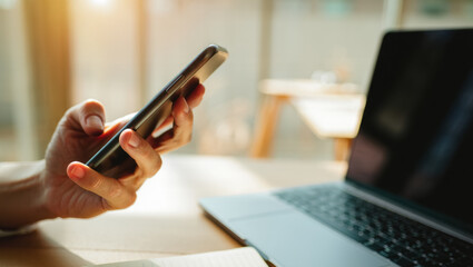 Fototapeta premium Close-up of a hand holding a smartphone beside a laptop on a desk in soft warm sunlight, creating a modern, calm and productive digital lifestyle atmosphere