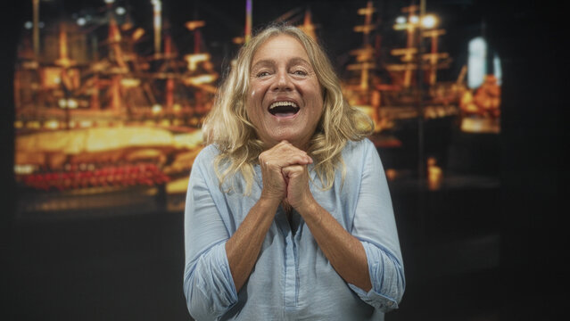 Senior woman clasping hands over chest, smiling with closed eyes in a studio with a lit ship exhibit and warm soft lighting; contentment.