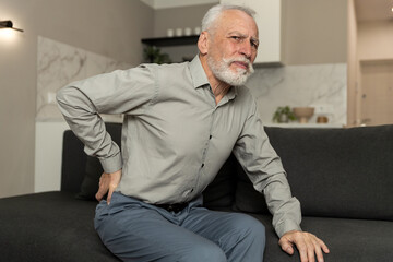 Fototapeta premium Elderly man suffering from lower back pain while sitting on sofa