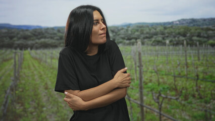 Naklejka premium Woman hugging her arms across her chest in a vineyard between trellis rows, hands clasped and looking sideways toward distant hills; quiet solitude.