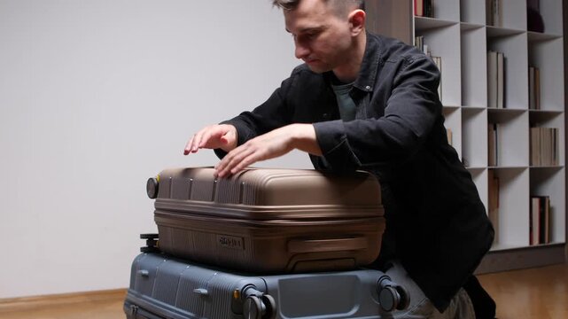 Man kneeling, pressing down on a full suitcase with both hands, attempting to close it for a trip while a stack of luggage sits ready for travel at home