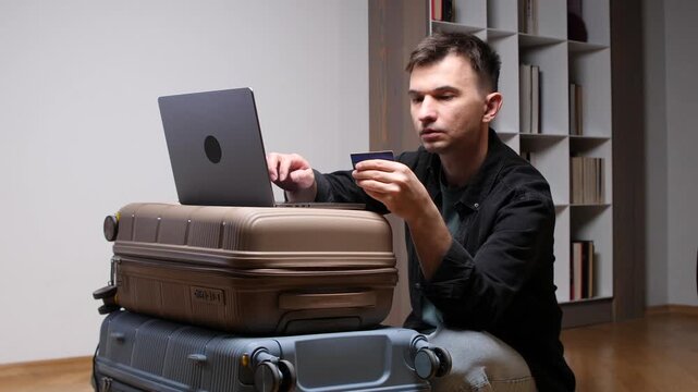 Man holding a blank credit card and typing on a laptop, making an online payment for a trip while sitting next to stacked luggage and planning a vacation