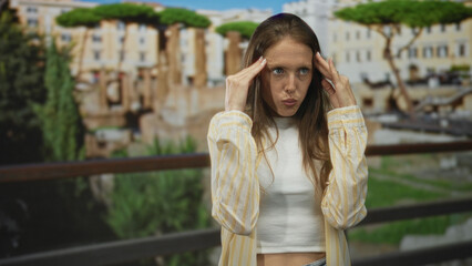 Fototapeta premium Woman touching temples with hands and mouth open at building among roman ruins outdoors; surprise wonder travel.