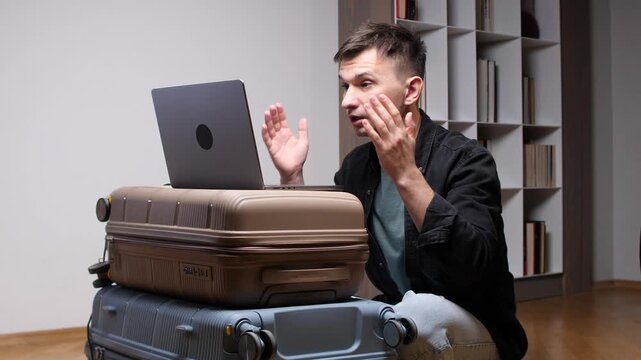 Man conducting a serious remote video conference from home, using a laptop perched on stacked suitcases while preparing for or returning from a business trip and gesturing during the call