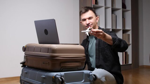 Man holding a small airplane model while planning international summer travel, sitting next to stacked luggage with a laptop on top, preparing for departure