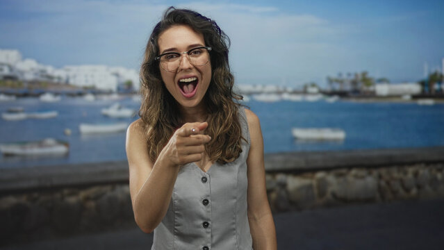 Young hispanic woman wearing glasses and a light grey vest points finger by sunlit seaside street; playful.