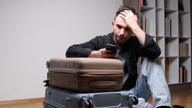 Stressed man sitting on the floor leaning on luggage, checking a smartphone with his hand on his head, dealing with travel issues or a cancelled flight